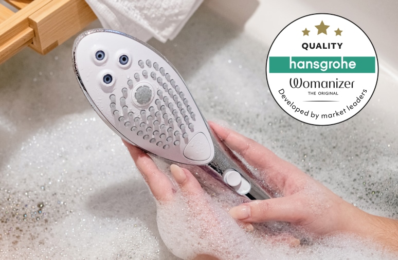 泡の入った浴槽で両手に持たれたシャワーヘッドと、右上に「hansgrohe」と「Womanizer」の品質ロゴ